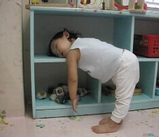 Un très jeune enfant, de deux ans environ, s'est endormi debout, la tête appuyée contre une étagère, comme foudroyé par le sommeil en plein jeu.